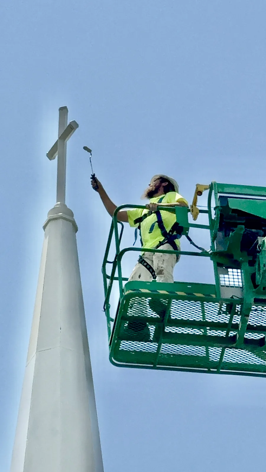 A harnessed ECHERER painter rolls fresh paint on a church steeple cross from a boom lift